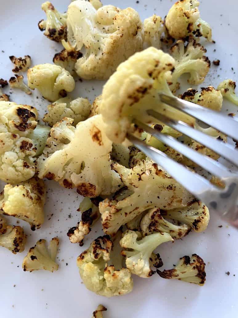 Close-up of a fork holding a roasted cauliflower floret over a plate of air-fried cauliflower.