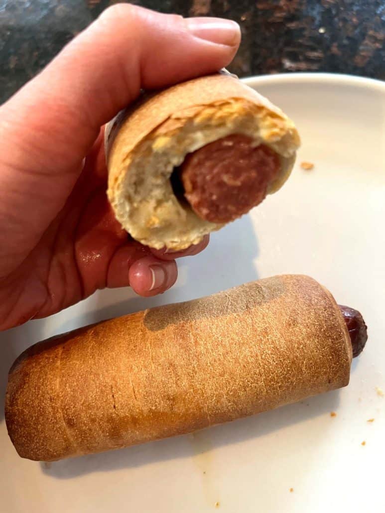 Melanie Mendelson from melaniecooks.com holding a bitten pretzel dog, showing the hot dog inside, with another whole pretzel dog on a plate in the background.