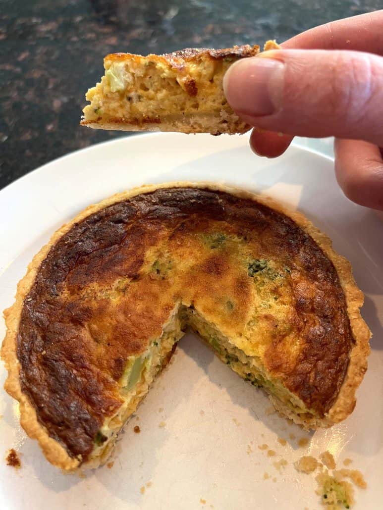 Melanie Mendelson from melaniecooks.com, holding a bite of broccoli and cheddar quiche above a partially sliced quiche on a plate, showing the soft filling and crisp crust from the air fryer method.