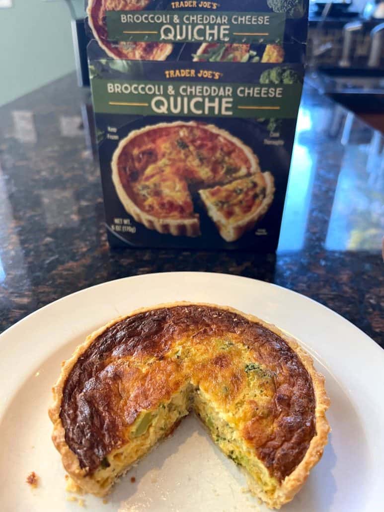 Cooked Trader Joe’s broccoli and cheddar quiche on a plate with a slice removed, shown in front of the product box to highlight results of the air fryer frozen quiche recipe.