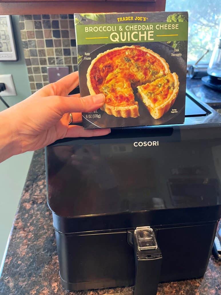 Melanie Mendelson from melaniecooks.com, holding Trader Joe’s frozen quiche box in front of an air fryer, ready to show how to cook it straight from frozen using the air fryer method.