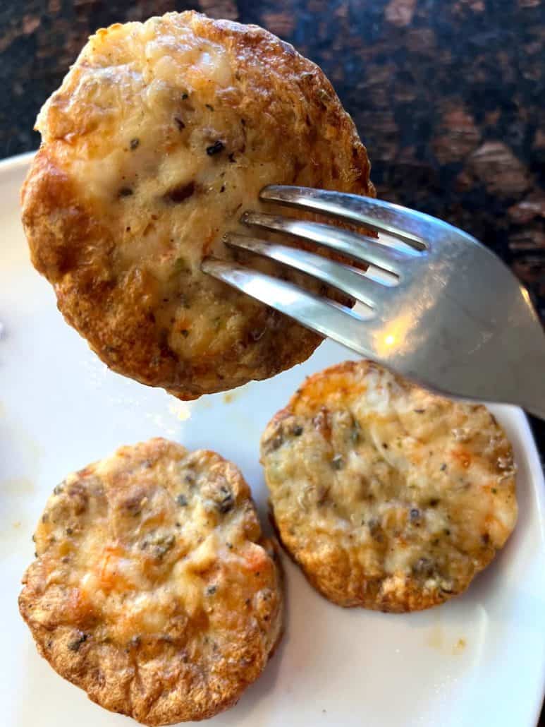 A crispy shrimp burger held by a fork, with two more shrimp burgers on a plate, freshly cooked and ready to eat.
