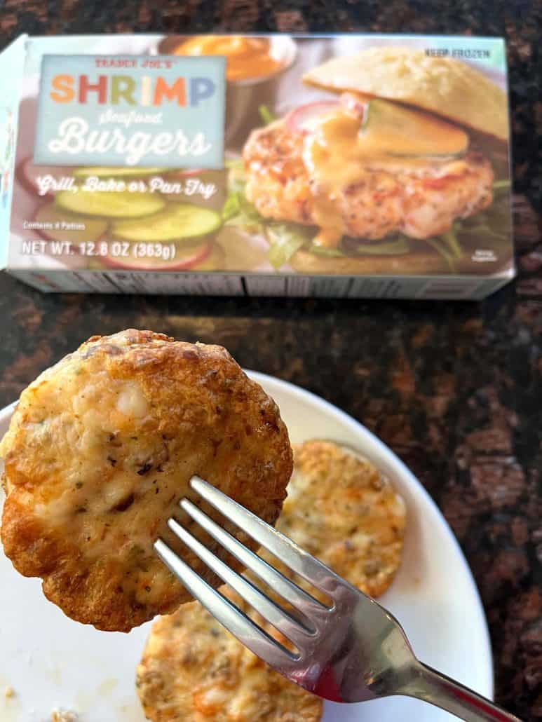 A crispy shrimp burger held by a fork, with more shrimp burgers on a plate and a Trader Joe’s Shrimp Burgers box in the background.