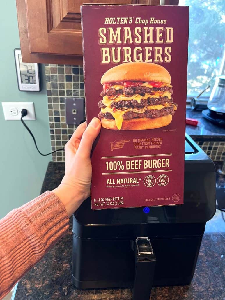 Melanie Mendelson from melaniecooks.com holding a box of frozen smashed burgers in front of the air fryer before cooking.