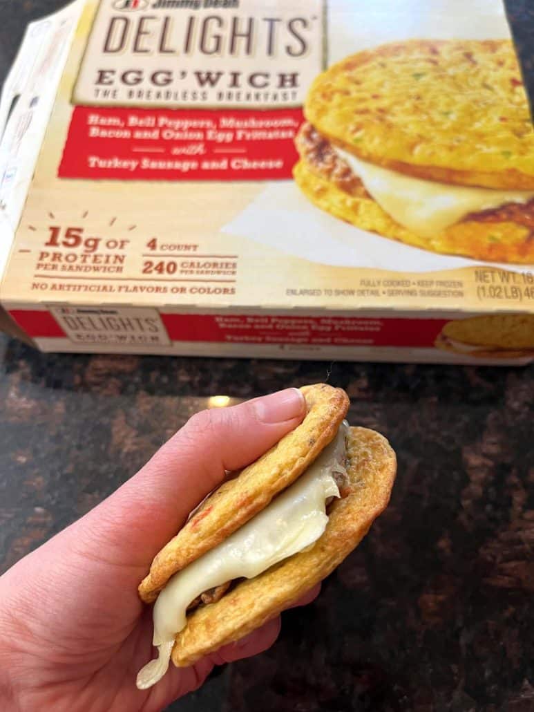 The hand of Melanie Mendelson from melaniecooks.com holding a freshly cooked Jimmy Dean Delights Egg’wich with melted cheese, in front of its packaging.