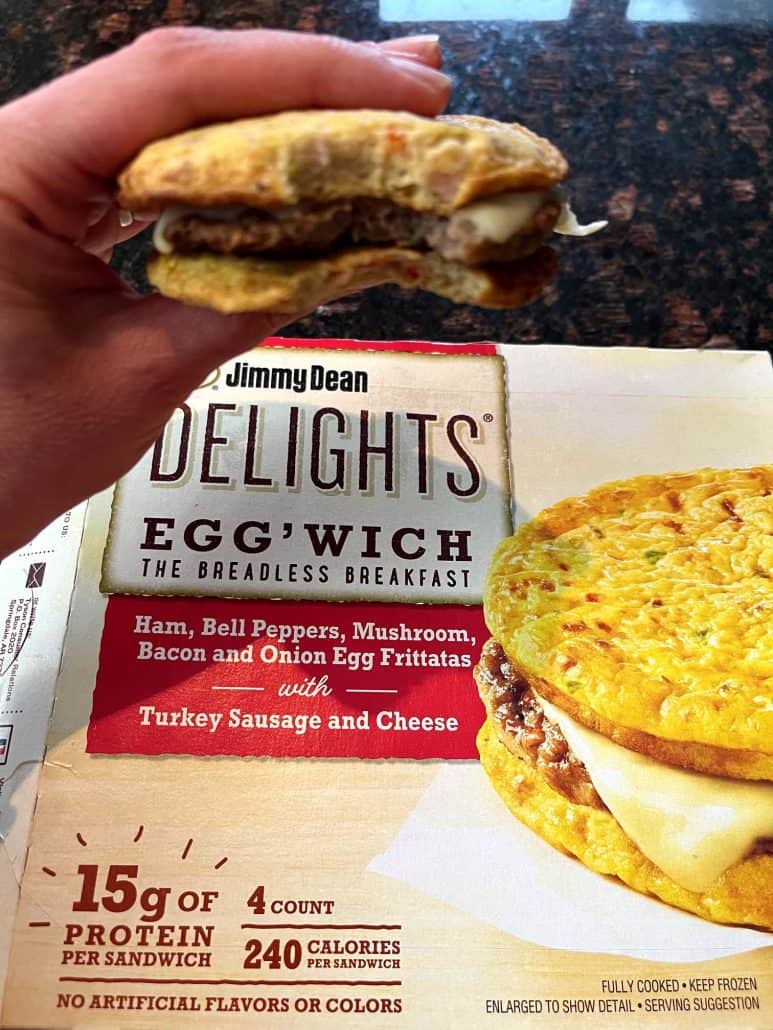 The hand of Melanie Mendelson from melaniecooks.com holding a bitten Jimmy Dean Delights Egg’wich in front of its packaging, showing the savory filling of this low-carb breakfast.