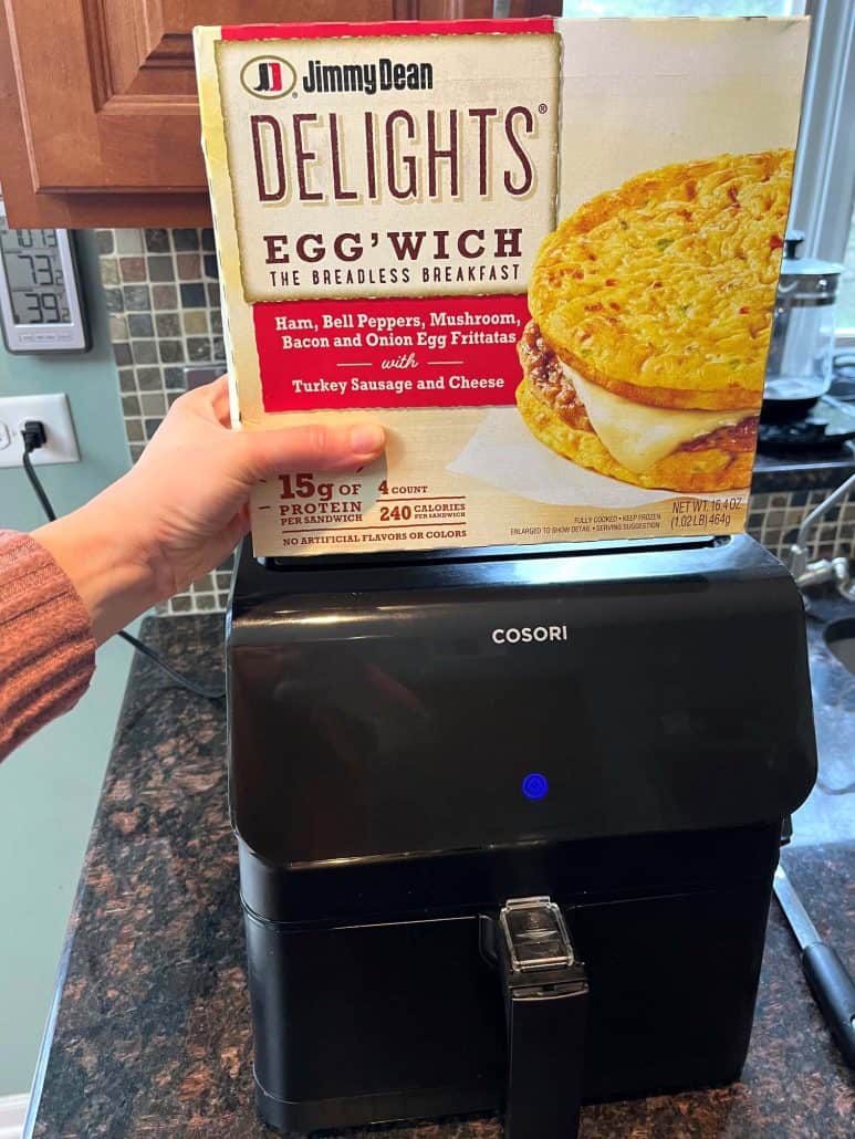 Hand of Melanie Mendelson from melaniecooks.com holding a box of Jimmy Dean Delights Egg’wich over an air fryer, ready to cook this low-carb breakfast sandwich to crispy perfection.