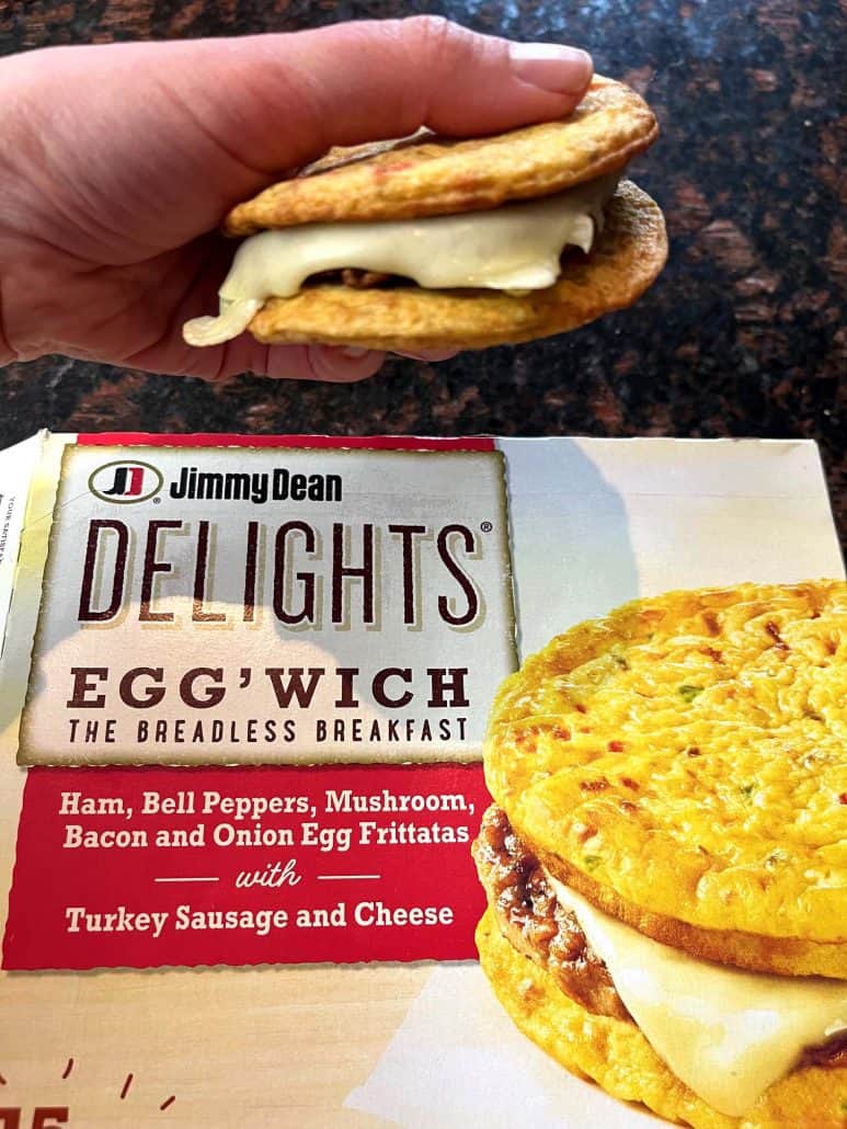 Hand of Melanie Mendelson from melaniecooks.com holding a delicious low-carb Jimmy Dean Delights Egg’wich, with gooey melted cheese, in front of its packaging.