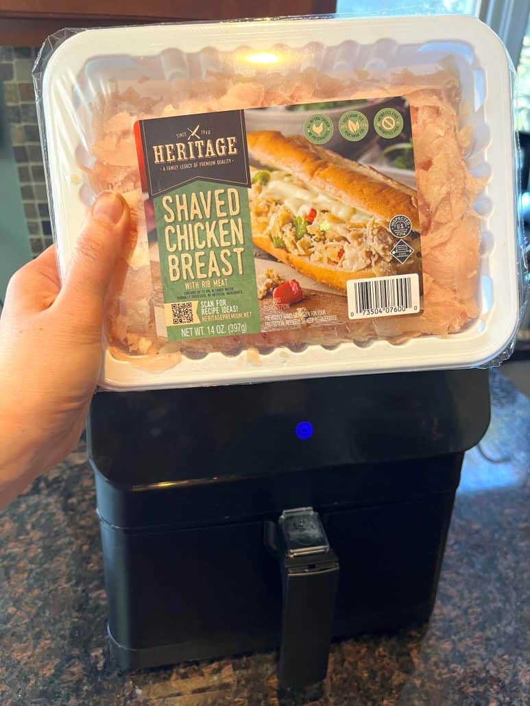 Hand of Melanie Mendelson from melaniecooks.com, holding a package of Kirkwood Shaved Chicken Breast in front of an air fryer—getting ready to cook.