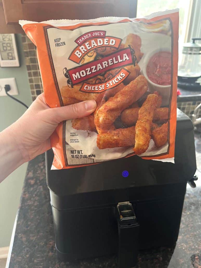 Melanie Mendelson from melaniecooks.com holding a bag of Trader Joe’s Breaded Mozzarella Sticks in front of an air fryer, ready to cook.