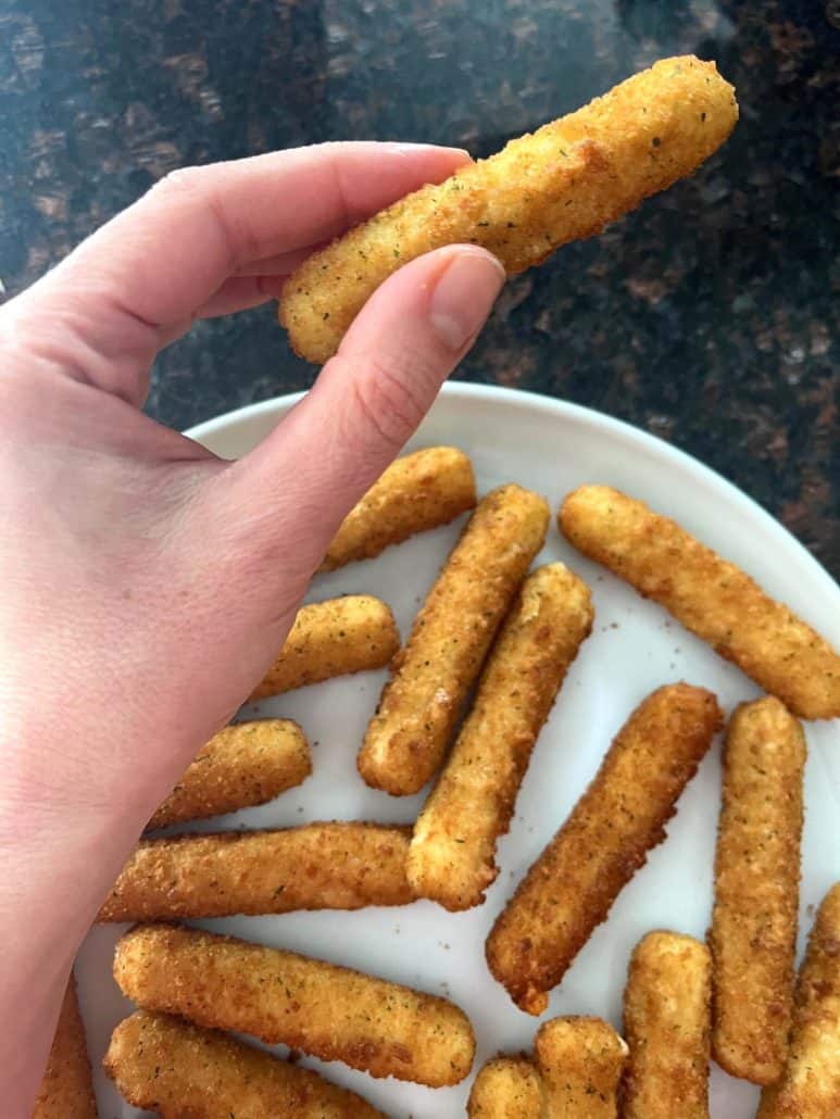 Melanie Mendelson from melaniecooks.com holding a crispy, golden mozzarella stick with a plate full of air-fried mozzarella sticks in the background.