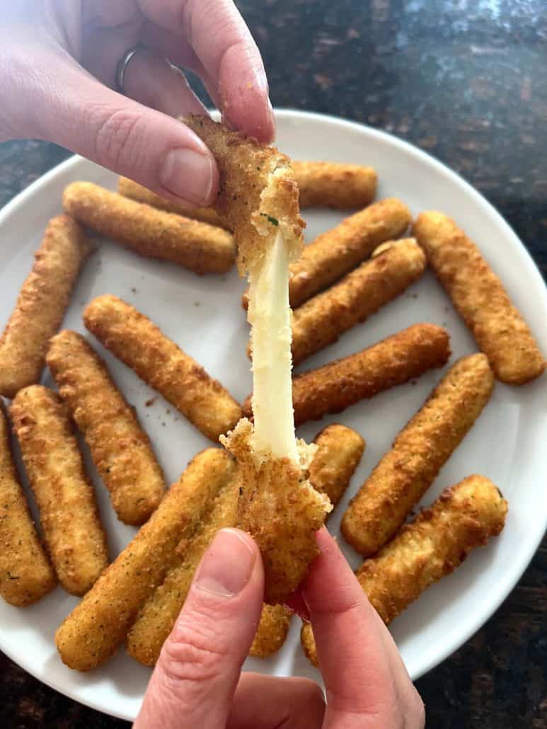 Melanie Mendelson from melaniecooks.com pulling apart a crispy mozzarella stick with gooey, melted cheese stretching between the halves, with a plate of air-fried mozzarella sticks in the background.
