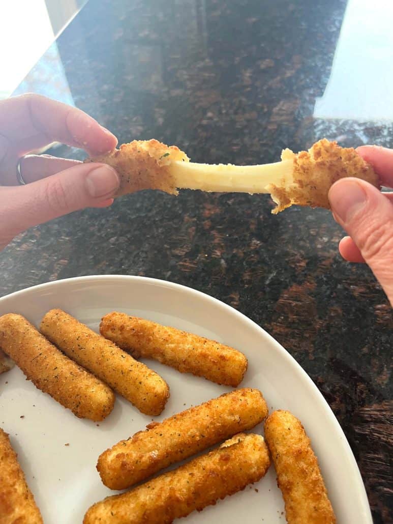 Melanie Mendelson from melaniecooks.com pulling apart a crispy mozzarella stick, revealing a stretchy, gooey melted cheese center, with a plate of air-fried mozzarella sticks in the background.