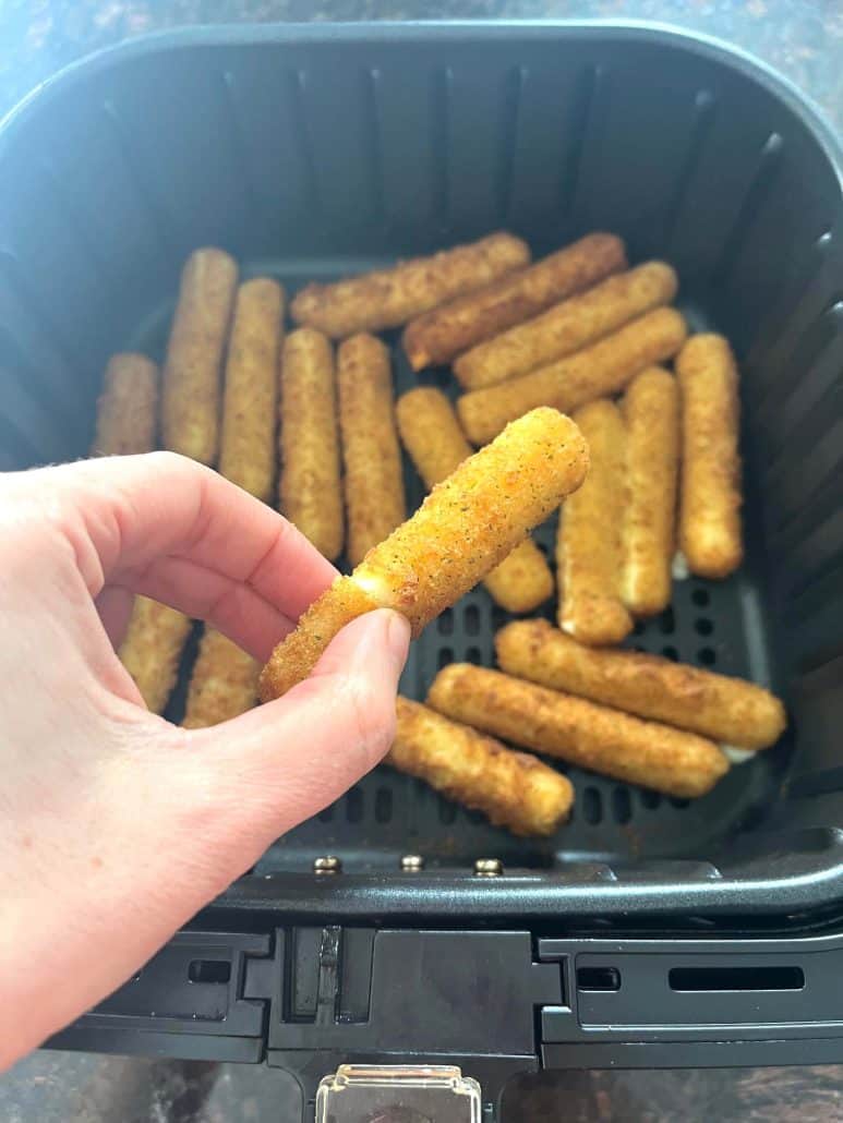 Melanie Mendelson from melaniecooks.com holding a crispy, golden mozzarella stick fresh from the air fryer, with more cooked mozzarella sticks in the basket.