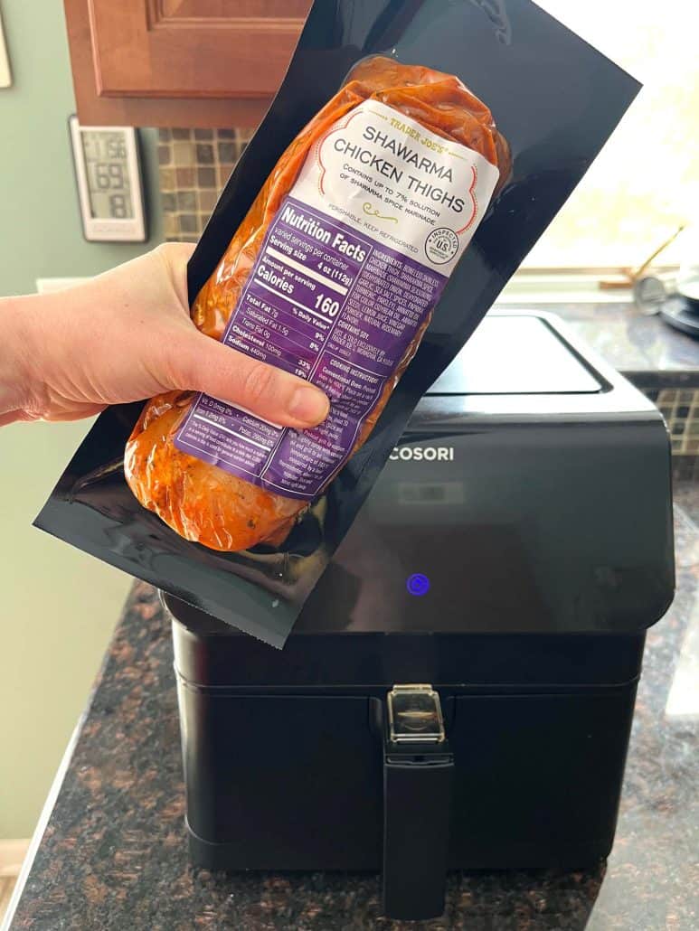 Melanie Mendelson from melaniecooks.com is holding a package of Trader Joe’s Shawarma Chicken Thighs in front of an air fryer, ready to cook for a quick and easy meal.