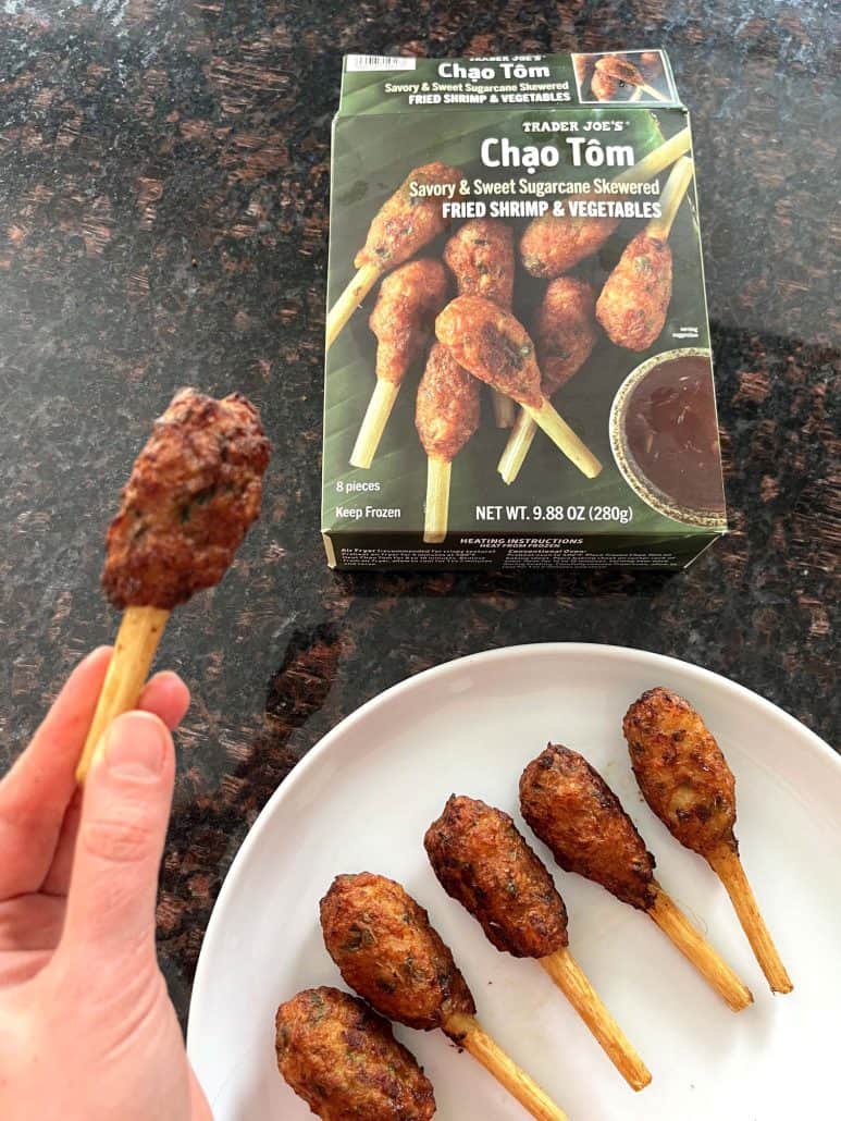 Hand of Melanie Mendelson from melaniecooks.com, holding a cooked Trader Joe’s Chao Tom with the box and a plate of finished skewers in the background.
