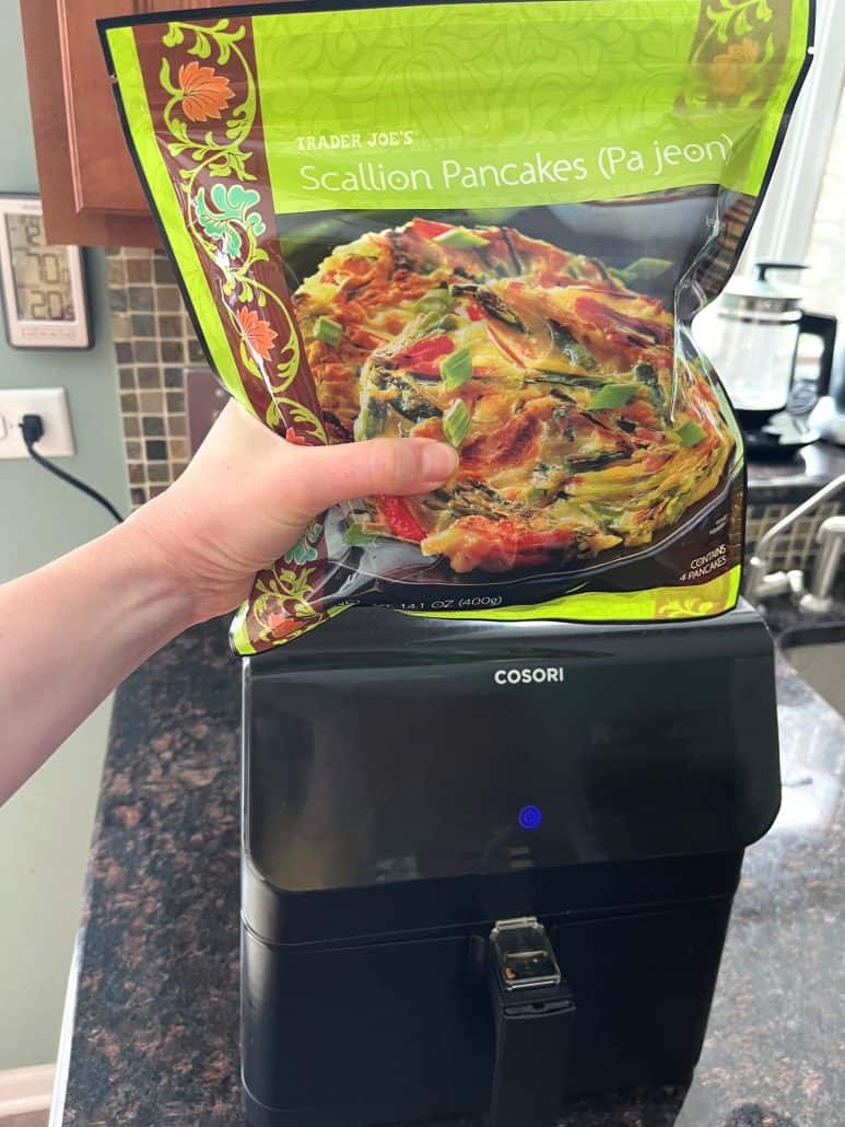 Melanie Mendelson from melaniecooks.com holding a bag of Trader Joe’s Scallion Pancakes in front of an air fryer, showing the product before cooking using the air fryer method.