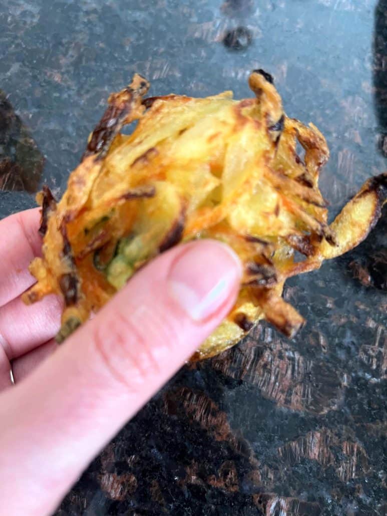 Melanie Mendelson from melaniecooks.com holding a crispy, golden-brown Trader Joe’s Vegetable Bird’s Nest, showing its crunchy tempura texture.