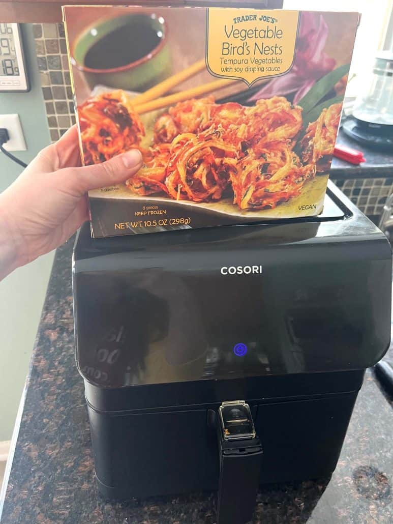 Melanie Mendelson from melaniecooks.com holding a box of Trader Joe’s Vegetable Bird’s Nests over an air fryer, ready to cook for a crispy and delicious appetizer.