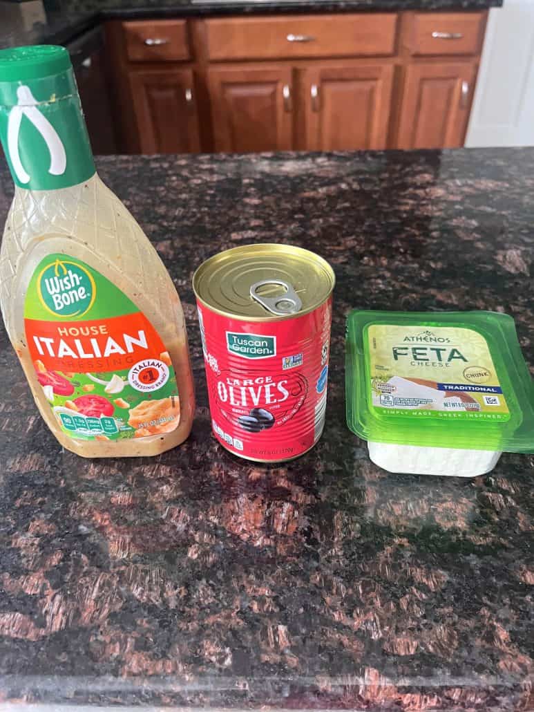 A bottle of Italian dressing, a can of black olives, and a container of feta cheese—simple ingredients for making marinated olives and feta cheese.