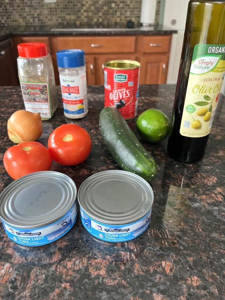 Fresh ingredients for Mediterranean Tuna Salad, including canned tuna, tomatoes, cucumber, onion, lime, olives, olive oil, and seasonings, laid out on a kitchen counter.