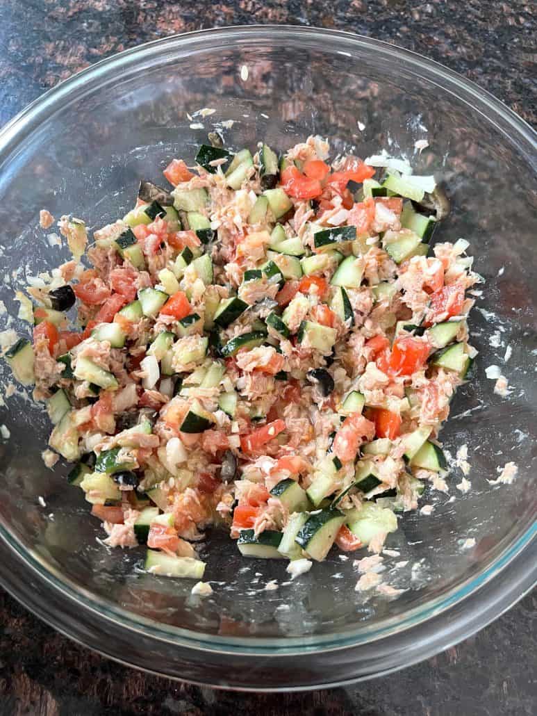 A glass bowl filled with freshly mixed Mediterranean Tuna Salad, featuring chopped cucumbers, tomatoes, olives, and tuna, tossed in olive oil and lime juice.