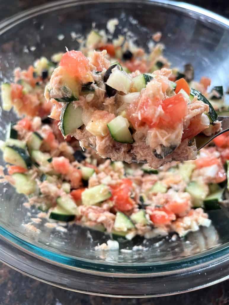 A spoonful of Mediterranean Tuna Salad lifted from a glass bowl, highlighting the fresh mix of tuna and other ingredients.