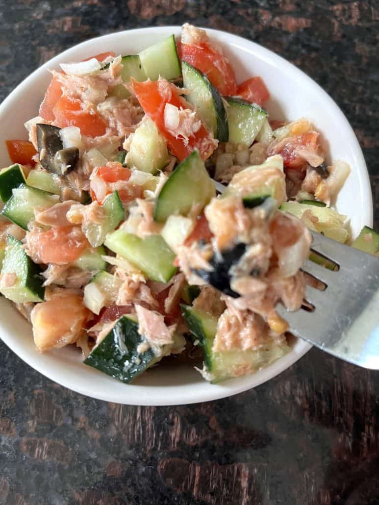 A forkful of Mediterranean Tuna Salad lifted from a white bowl, showcasing fresh tuna, cucumbers, tomatoes, olives, and a light olive oil and lime dressing.