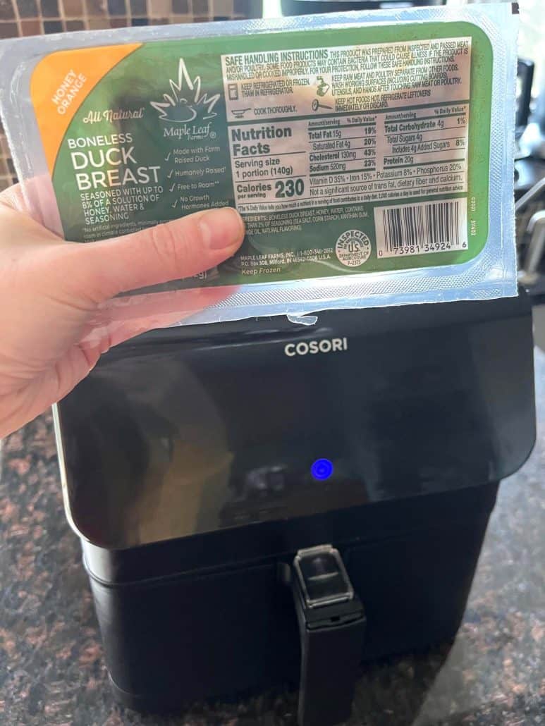 Melanie Mendelson from melaniecooks.com, holding a package of frozen duck breast in front of an air fryer, showing the ingredient before making the Air Fryer Duck Breast recipe.