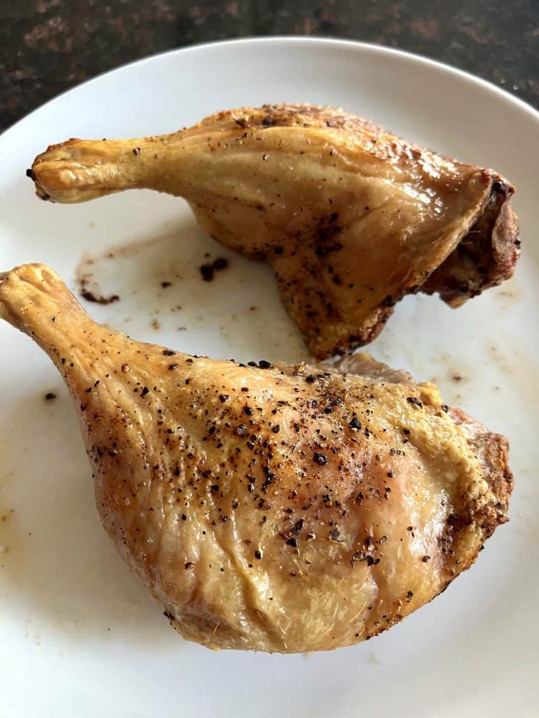 Side view of two crispy duck legs on a white plate, showing the juicy texture and golden skin.