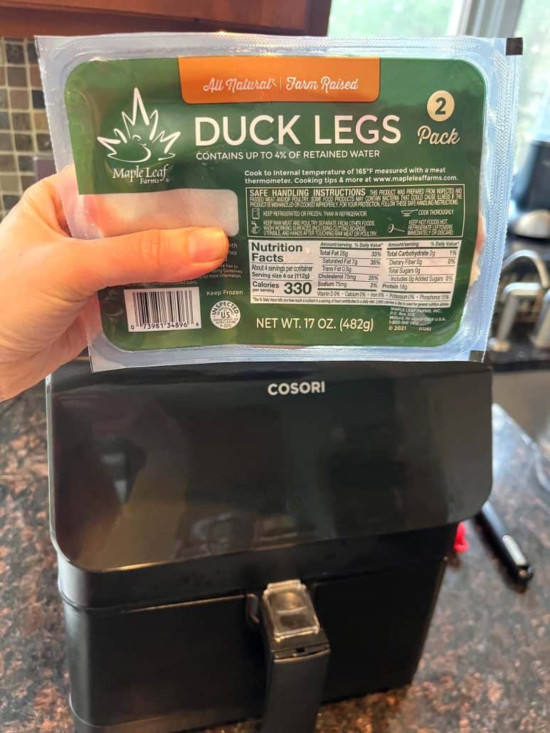 Melanie Mendelson from melaniecooks.com holding a package of duck legs in front of an air fryer, ready to start cooking the Air Fryer Duck Legs recipe.