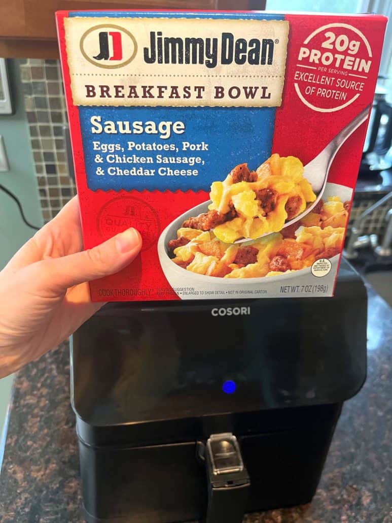 Melanie Mendelson from melaniecooks.com, holding a Jimmy Dean Sausage Breakfast Bowl box in front of an air fryer—getting ready to cook. 