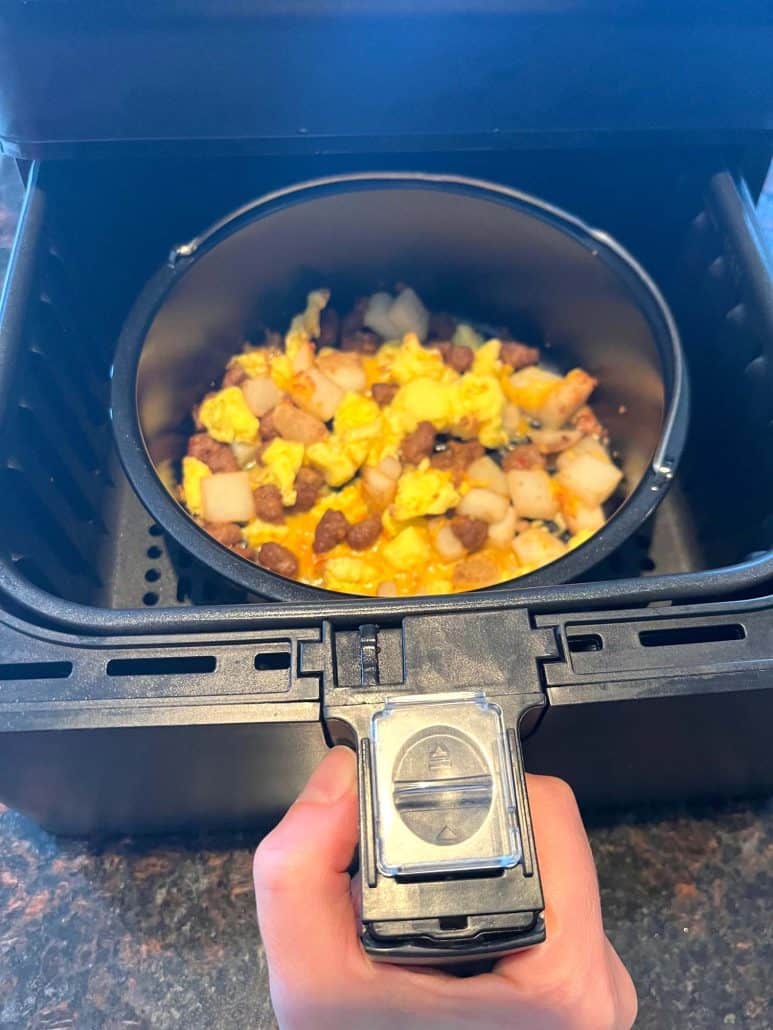 Melanie Mendelson from melaniecooks.com, pulling out air fryer basket with a cooked Jimmy Dean breakfast bowl inside, showing melted cheese on top.