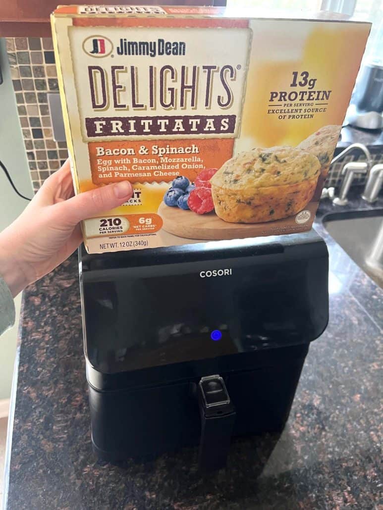 The hand of Melanie Mendelson from melaniecooks.com holding a box of Jimmy Dean Delights Frittatas in front of an air fryer.