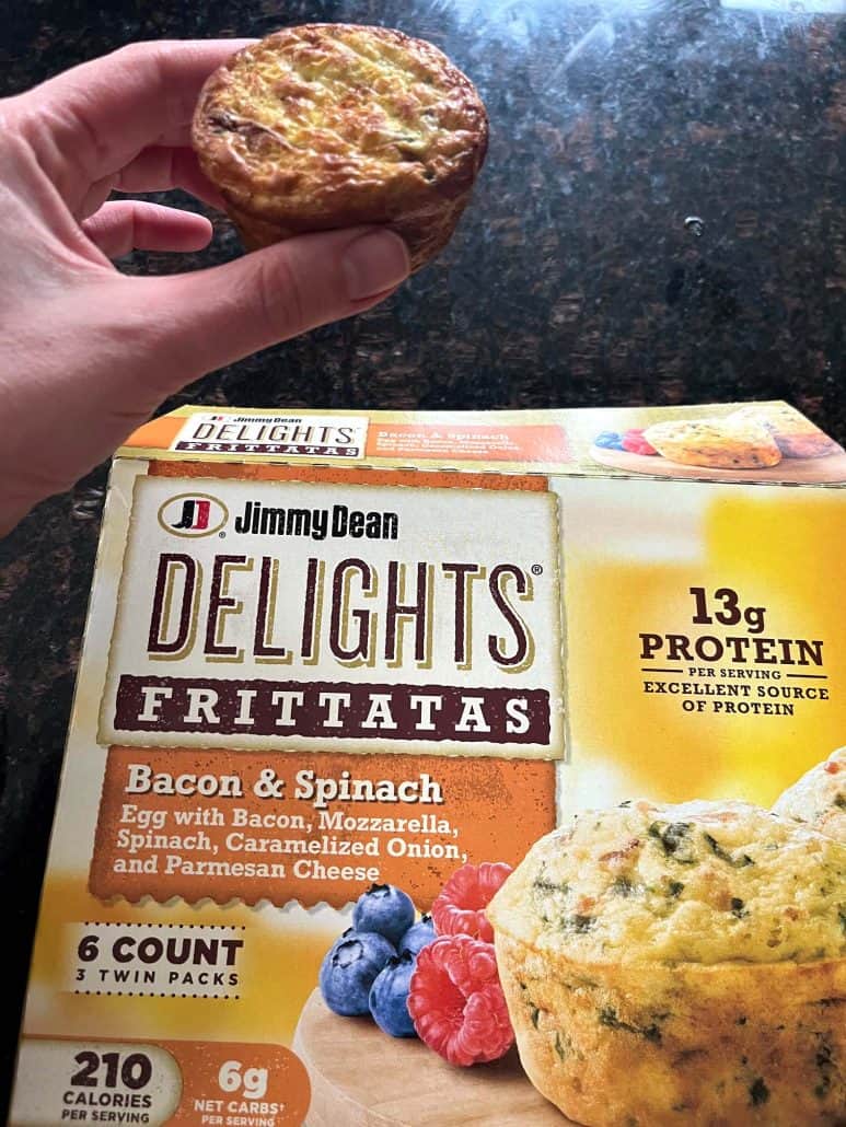 The hand of Melanie Mendelson from melaniecooks.com holding a cooked Jimmy Dean Bacon & Spinach Frittata in front of the product box, showing the ready-to-eat result of air frying straight from frozen.