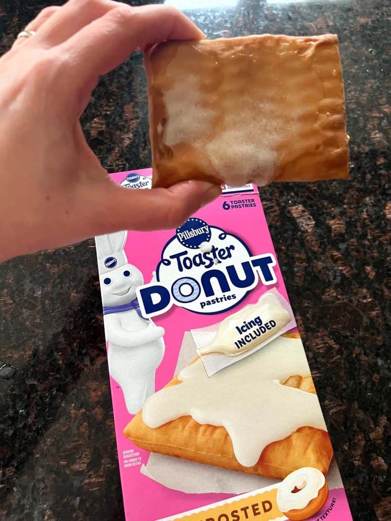 Melanie Mendelson from melaniecooks.com is holding a frosted Pillsbury Toaster Donut Pastry above the product box, showing the finished air fryer pastry next to its packaging.