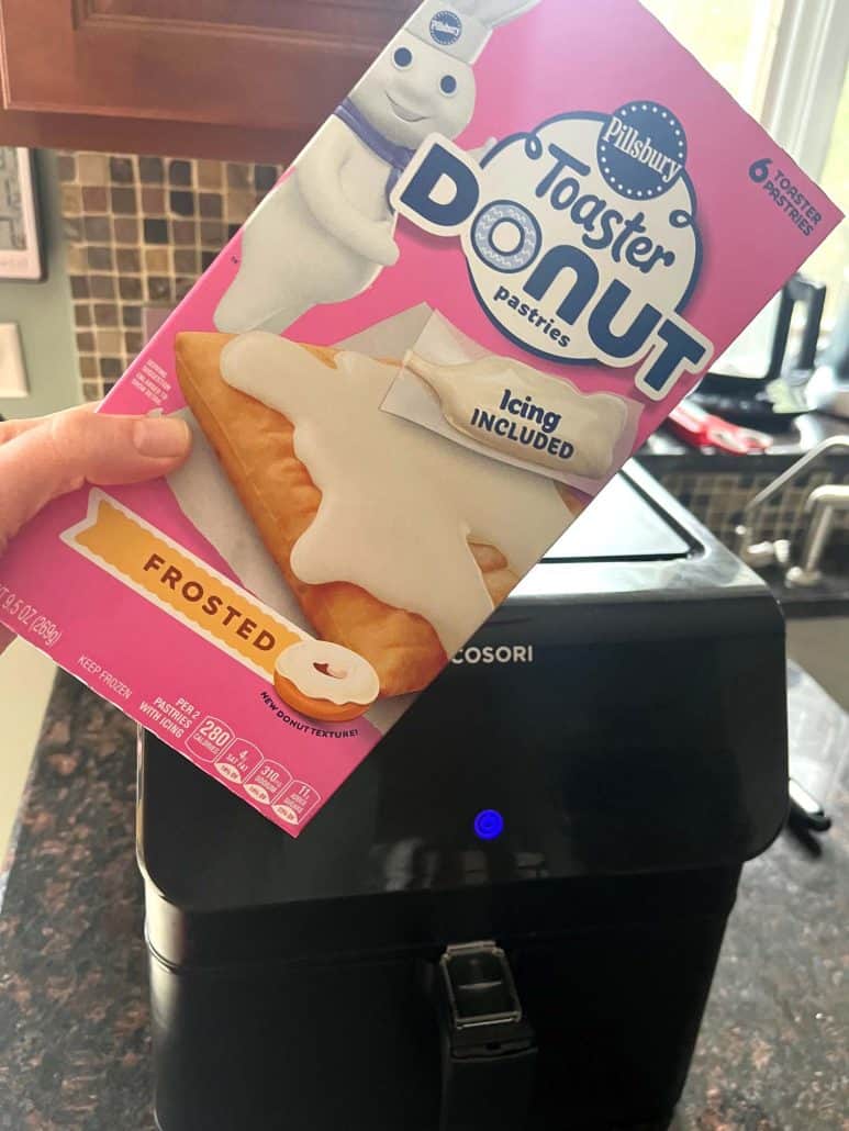 Melanie Mendelson from melaniecooks.com holding a box of Pillsbury Toaster Donut Pastries in front of an air fryer, showing the product used to make easy air fryer toaster donuts.