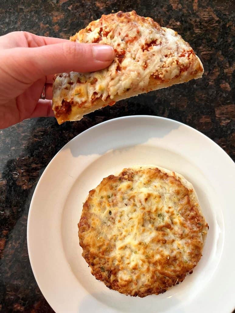 Melanie Mendelson from melaniecooks.com holding a slice of Trader Joe’s Bambino Pizza to show melted cheese, with another whole pizza on a white plate below.