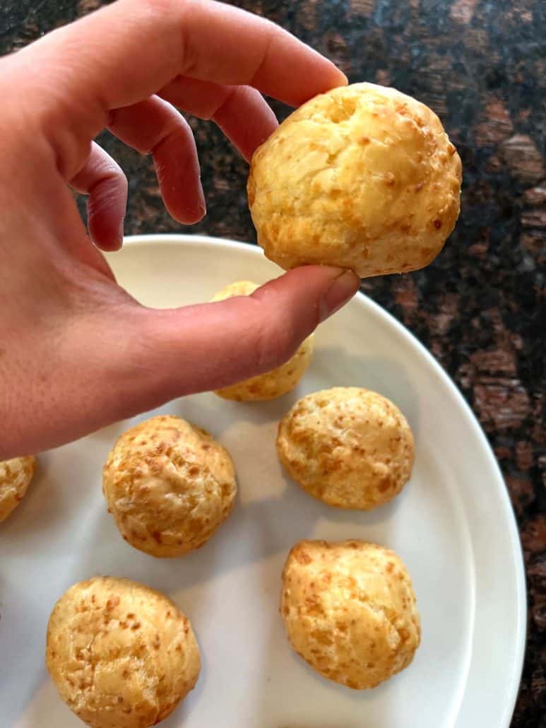 Melanie Mendelson from melaniecooks.com is holding a piece of Brazilian cheese bread over a plate. Showing the light, fluffy texture of this easy air fryer appetizer.