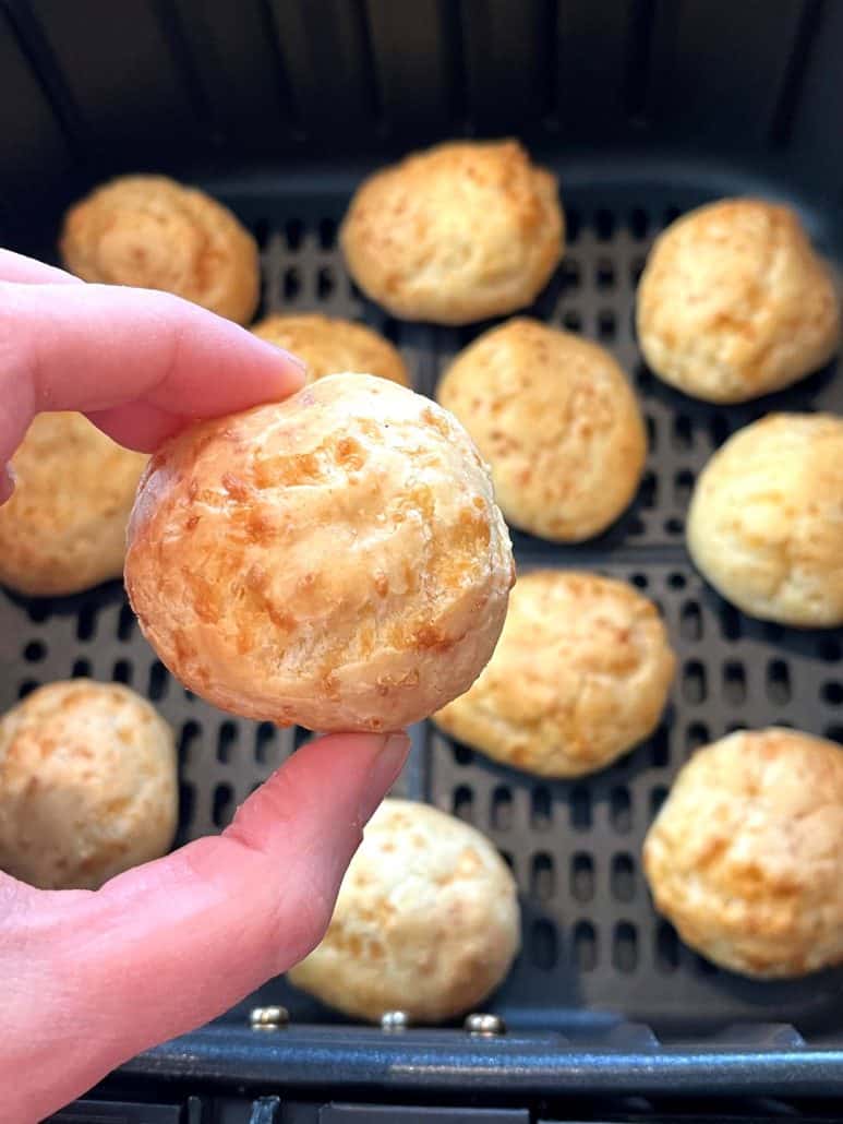 Melanie Mendelson from melaniecooks.com holding a piece of golden brown Brazilian cheese bread in front of an air fryer basket—showing the perfect texture of this easy air fryer snack.