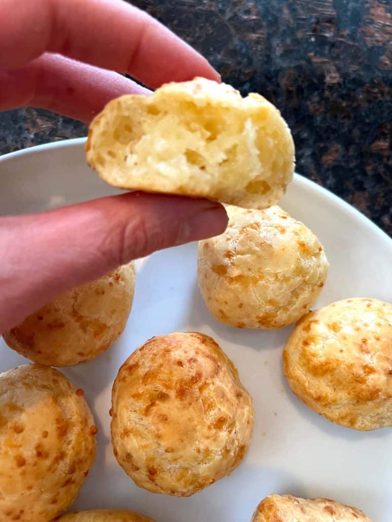 Melanie Mendelson from melaniecooks.com holding a Brazilian cheese bread with a bite taken out, showing the soft and cheesy inside—perfect result from this easy air fryer recipe.