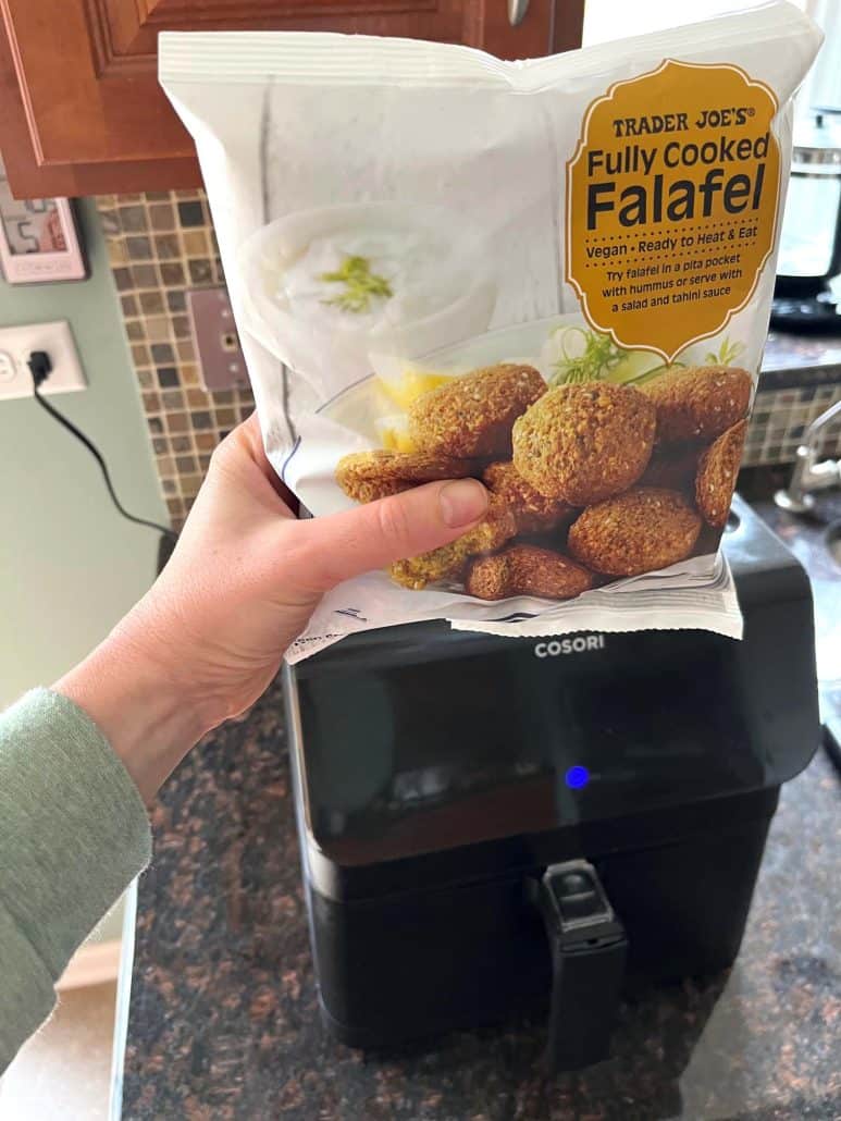 Melanie Mendelson from melaniecooks.com is holding a bag of Trader Joe’s Frozen Falafel next to an air fryer, ready to make quick, crispy falafel.