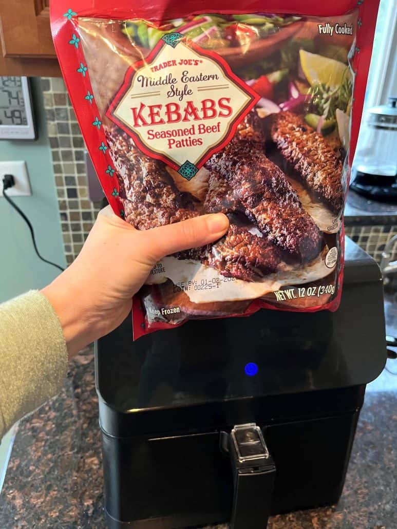 Melanie Mendelson from melaniecooks.com holding a package of Trader Joe’s frozen beef kebabs in front of an air fryer, ready to show how to cook them quickly and easily.