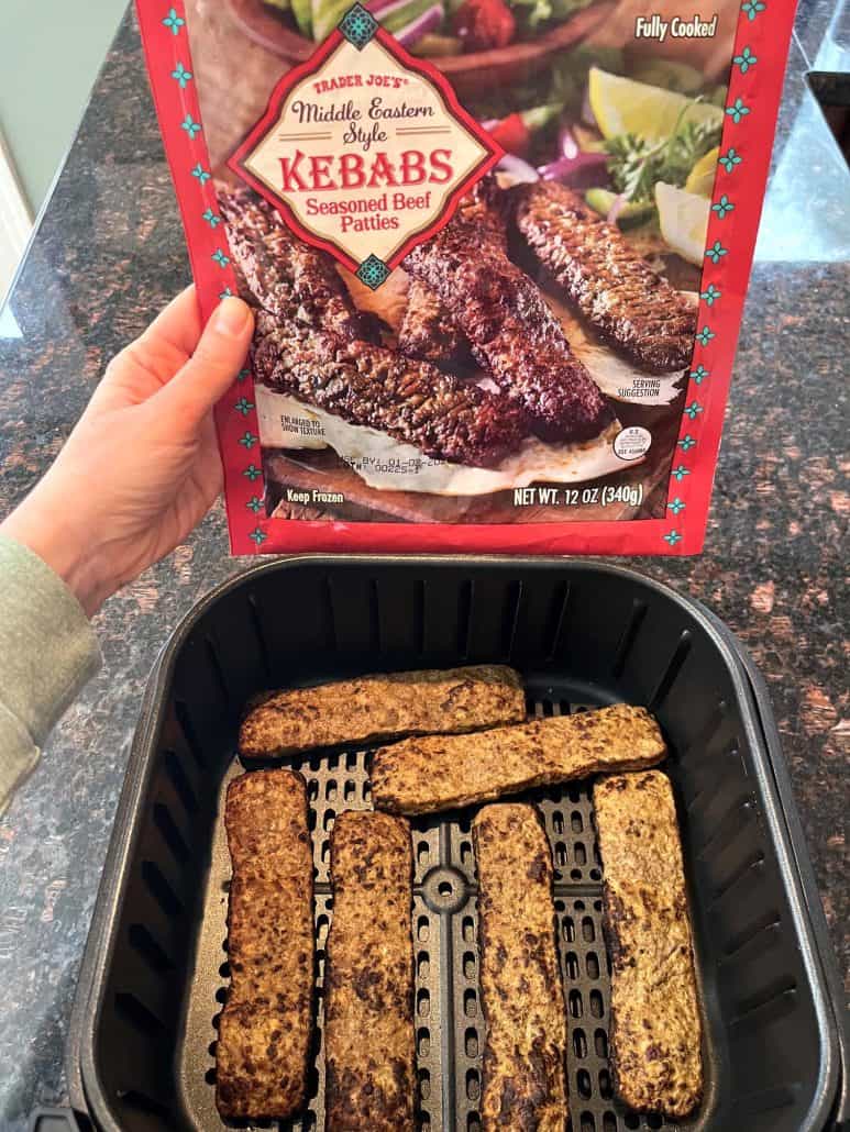 Melanie Mendelson from melaniecooks.com holding Trader Joe’s frozen beef kabobs package over an air fryer basket filled with cooked kabobs, showing the before-and-after of this easy air fryer recipe.
