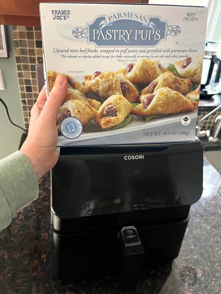 Hand of Melanie Mendelson from melaniecooks.com, holding a box of Trader Joe's Pastry Pups in front of an air fryer, ready to cook for a quick and crispy appetizer.