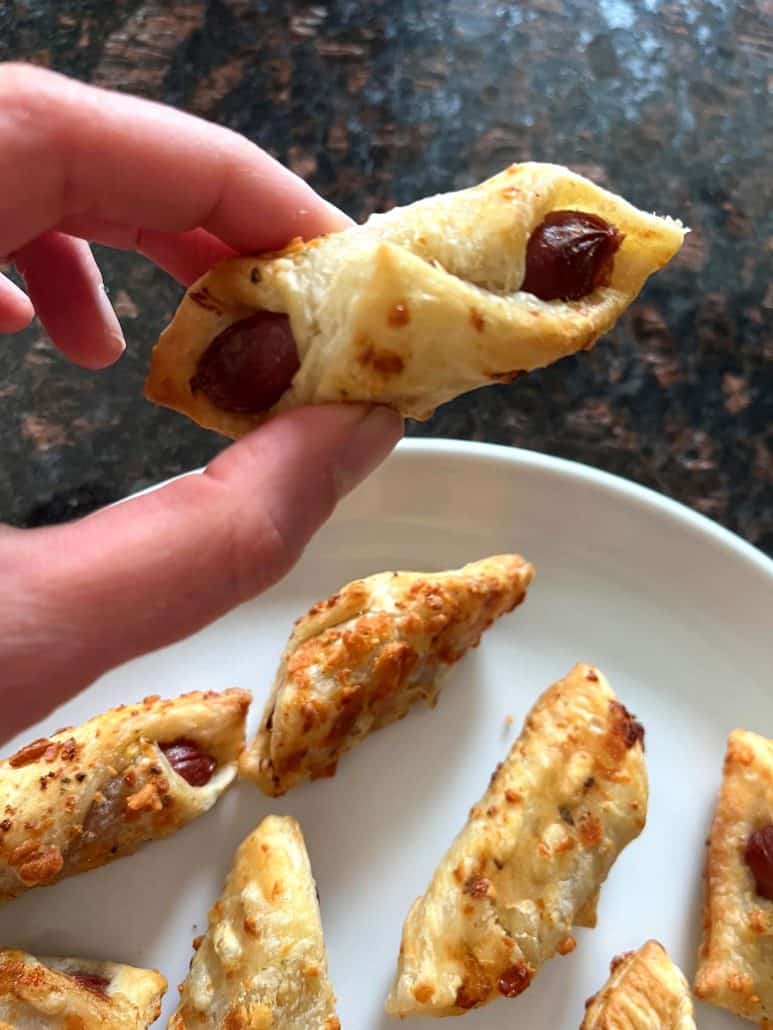 Hand of Melanie Mendelson from melaniecooks.com, holding a crispy Trader Joe's Pastry Pup over a plate of cooked pastry pups, showing the flaky golden pastry wrapped around the mini hot dog.
