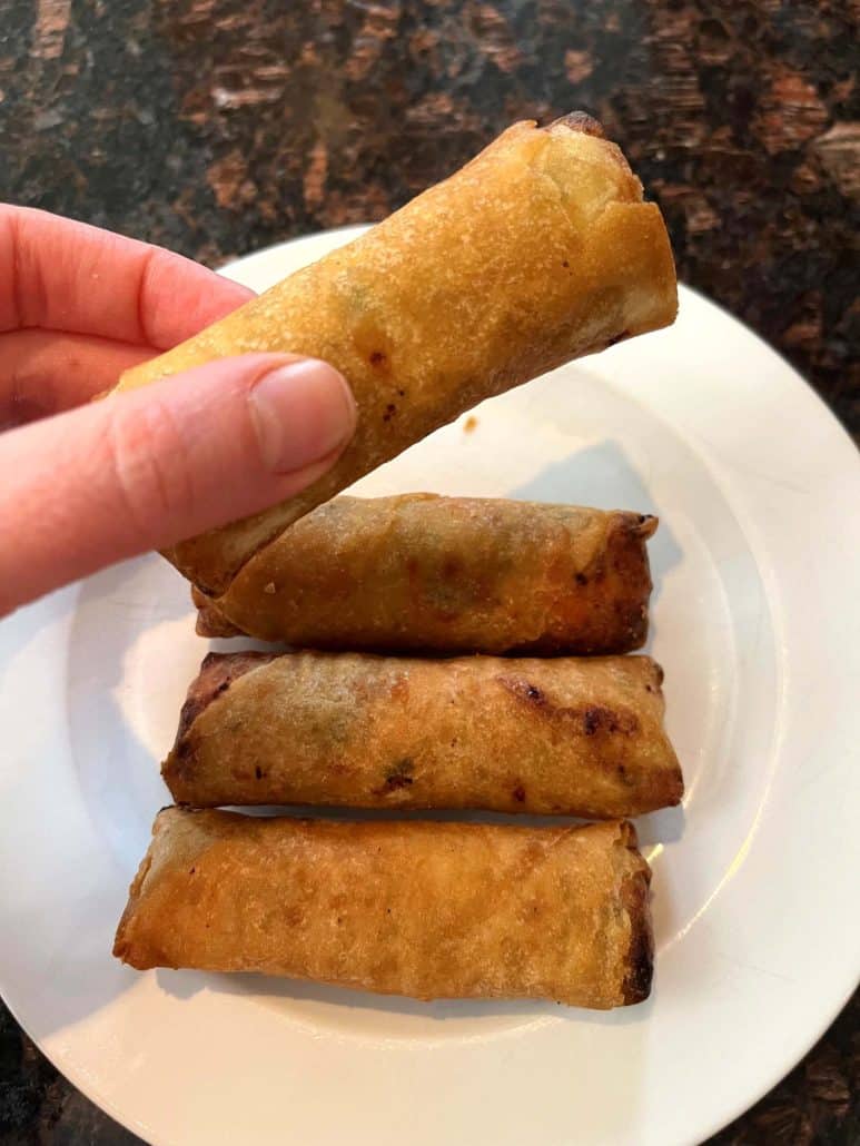 Melanie Mendelson from melaniecooks.com holding a crispy Trader Joe’s Chicken Spring Roll over a plate with more spring rolls, ready to enjoy.