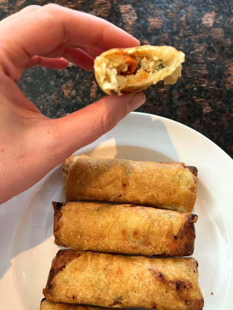 Melanie Mendelson from melaniecooks.com holding a Trader Joe’s Chicken Spring Roll with a bite taken out, showing the juicy filling—served over a plate of crispy air-fried spring rolls.