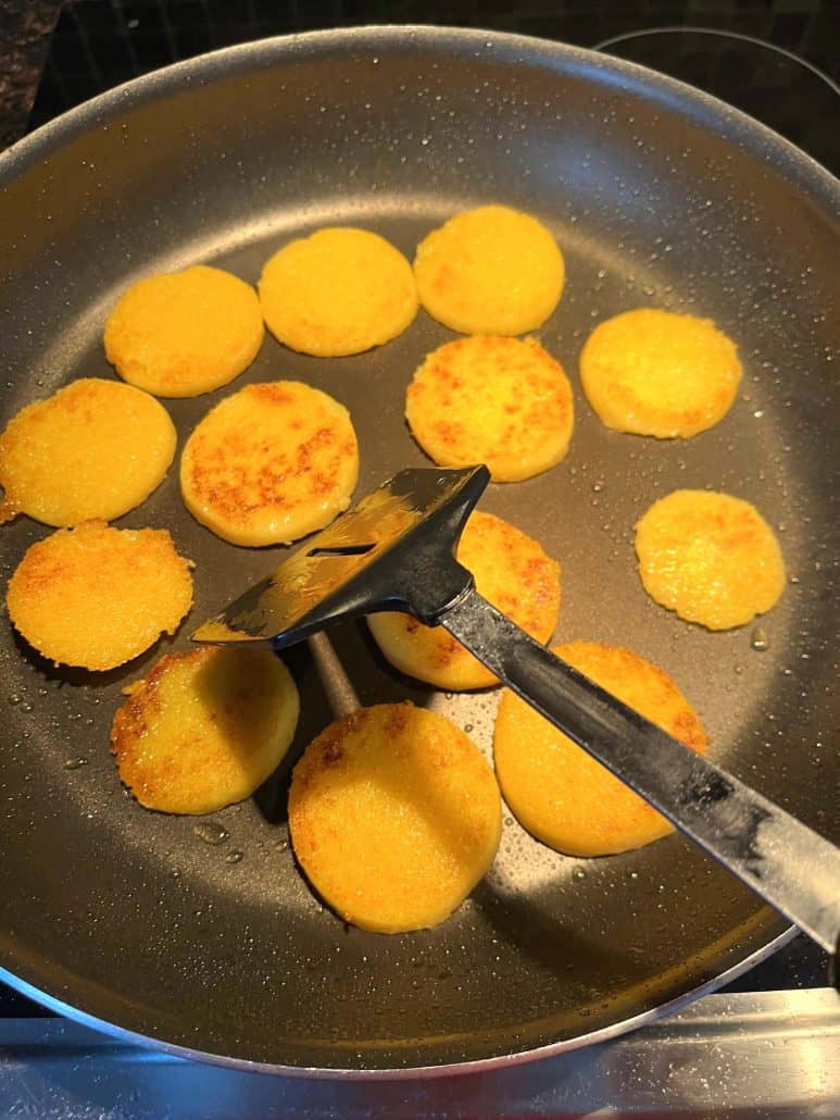 Using tongs to flip golden brown polenta slices in a frying pan, cooking them to crispy perfection.