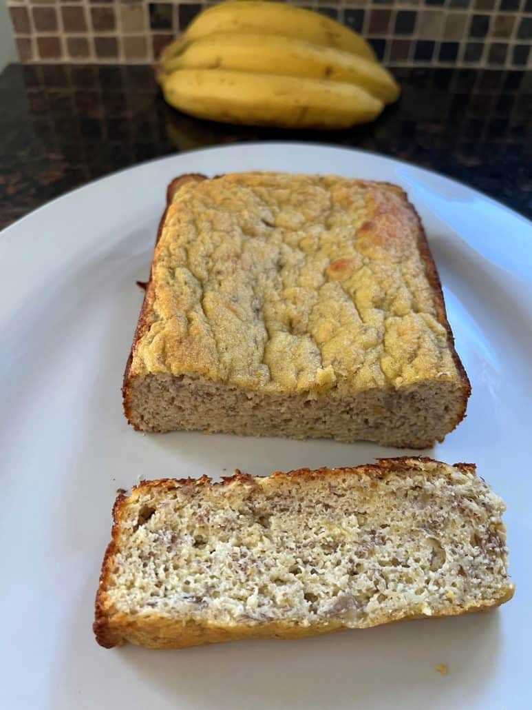 Moist slice of almond flour banana bread on a white plate with ripe bananas in the background.